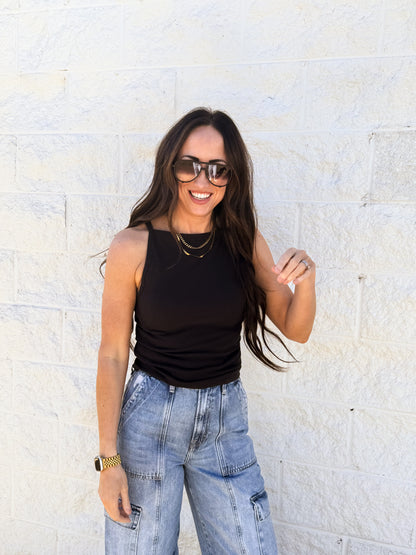 Woman wearing a black tank top and blue jeans standing against a light stone wall.