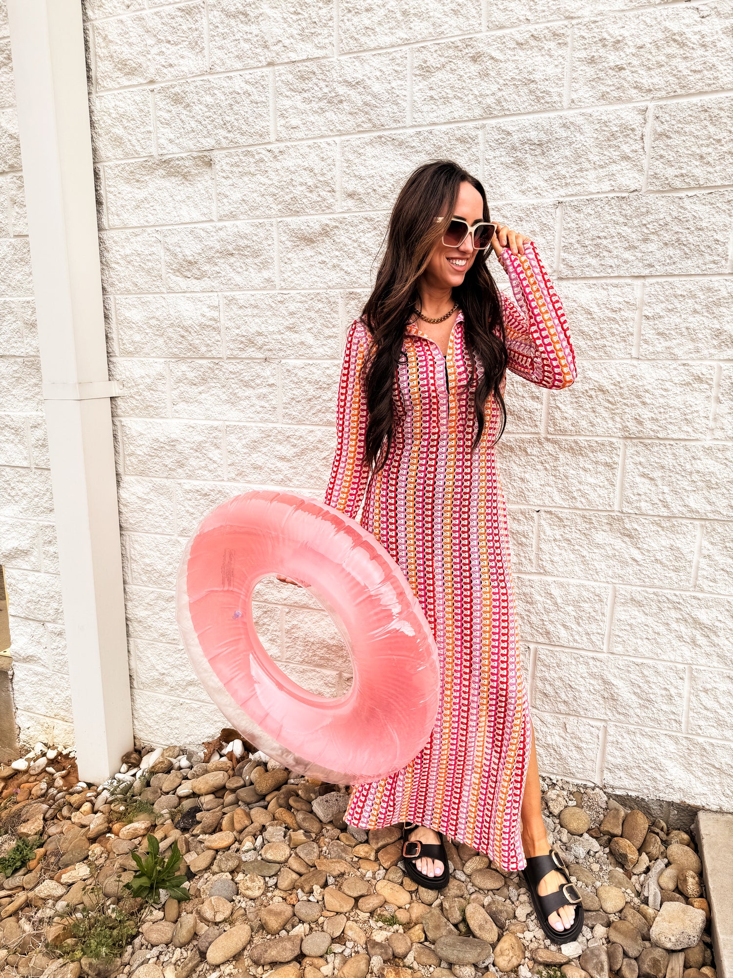 Woman wearing a red and pink patterned long sleeve maxi dress styled with black buckle sandals and oversized sunglasses, holding a pink pool float, standing against a white brick wall for a fun spring outfit look.