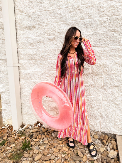 Woman wearing a red and pink patterned long sleeve maxi dress styled with black buckle sandals and oversized sunglasses, holding a pink pool float, standing against a white brick wall for a fun spring outfit look.