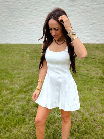 A woman standing in an open field wearing a white mini dress with a square neckline and pleated design.