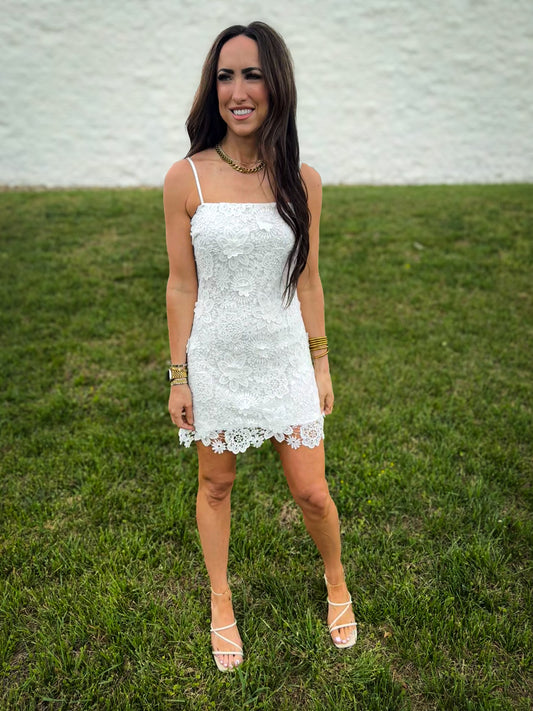 Woman wearing a white floral lace spaghetti-strap mini dress with a scalloped hem, styled with gold jewelry and strappy heels, standing on grass in front of a light textured wall.