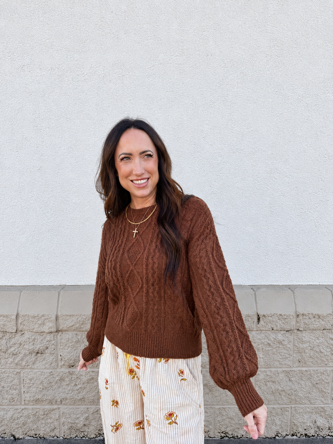 Model wearing the Cable Knit Balloon Sleeve Sweater in Chestnut, featuring a rich brown color, textured cable-knit design, relaxed fit, crew neckline, voluminous balloon sleeves, and ribbed cuffs and hem, styled with ivory floral drawstring pants.
