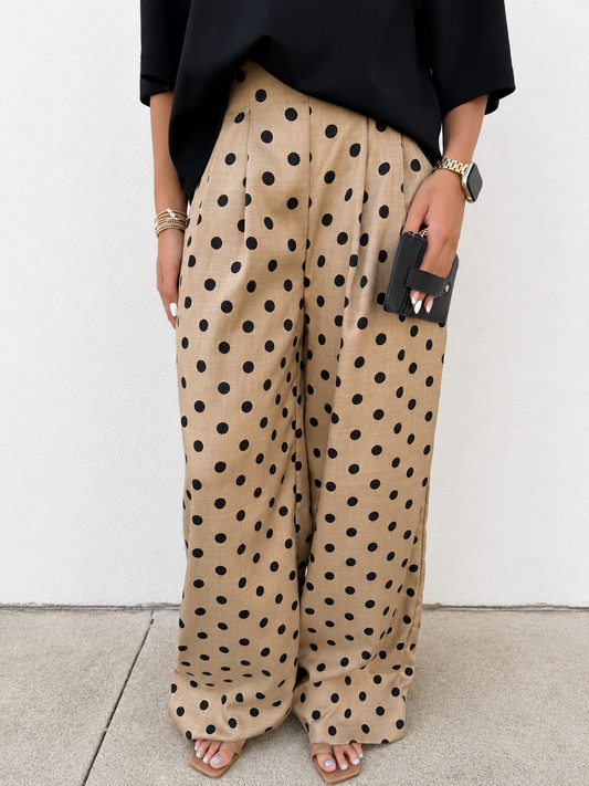 A person wearing beige wide-leg pants with a black polka dot pattern, styled with a black top and sandals, standing against a white wall.