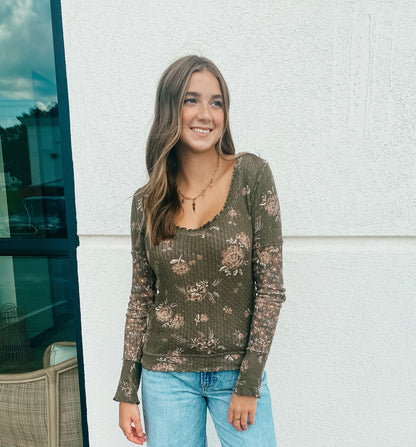 Woman wearing an army green, floral long-sleeve top and jeans standing against a white wall.