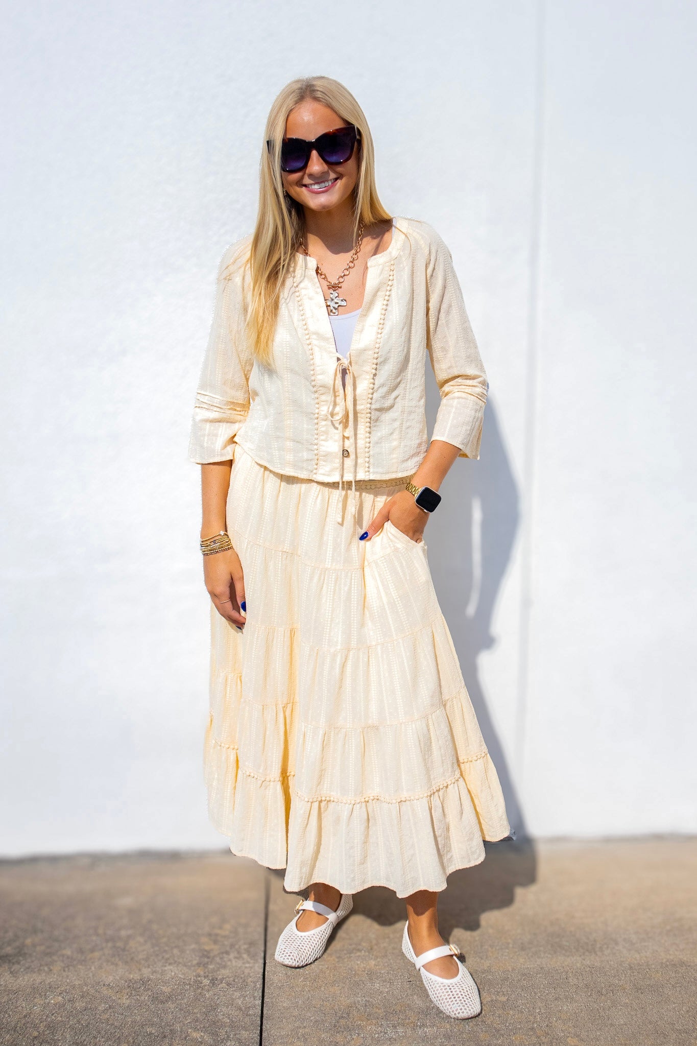 A woman wearing a cream-colored two-piece outfit consisting of a long-sleeve button-up shirt and a matching tiered maxi skirt, styled with white flat shoes and sunglasses against a white wall.