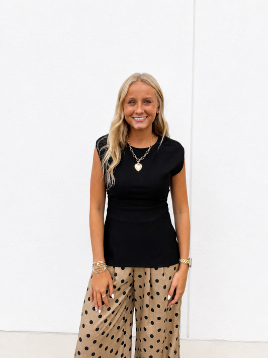 A smiling blonde woman wearing a black short-sleeve top and beige wide-leg pants with black polka dots, standing against a plain white wall.