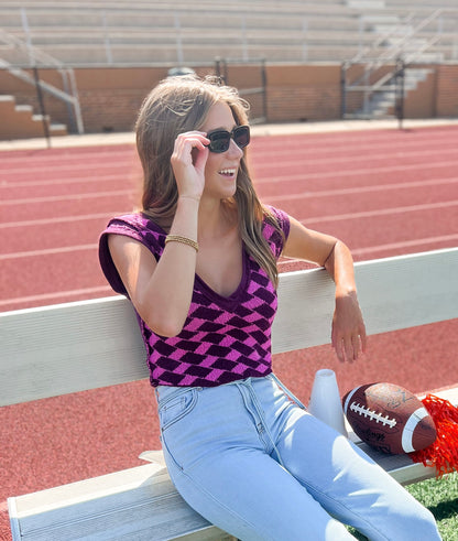 Woman sitting on a bench at a stadium with a football and pom-pom nearby wearing a purple and pink checkered, v-neck sweater and light denim.