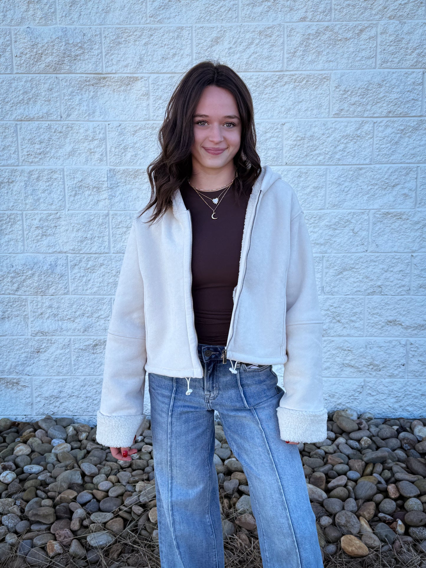 Person wearing a white jacket, brown top, and blue jeans standing against a light gray brick wall.