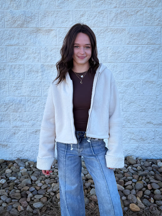 Person wearing a white jacket, brown top, and blue jeans standing against a light gray brick wall.