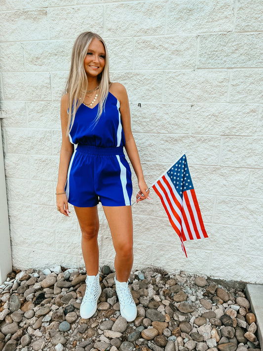 A woman standing on a rocky surface holding an American flag, wearing a royal blue romper with white vertical stripes, an elastic waistband, and pockets, paired with white sneakers.