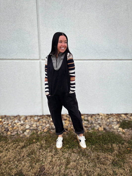 Woman wearing a black casual jumpsuit layered over a striped long sleeve top with white sneakers, styled in a relaxed outfit outdoors.