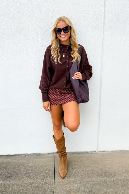 A woman posing in front of a white wall wearing a dark brown button-up shirt, a red and black checkered mini skirt, brown suede knee-high boots, and holding a dark purple tote bag.