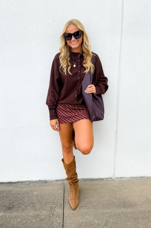 A woman posing in front of a white wall wearing a dark brown button-up shirt, a red and black checkered mini skirt, brown suede knee-high boots, and holding a dark purple tote bag.