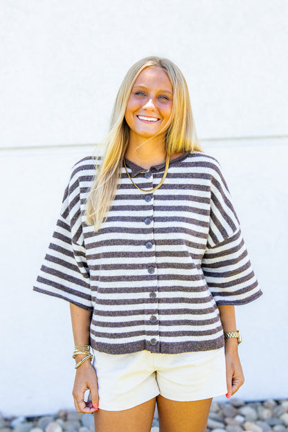 A smiling blonde woman wearing a brown and white horizontally striped button-up cardigan with elbow-length sleeves, paired with cream shorts, standing against a white stucco wall.