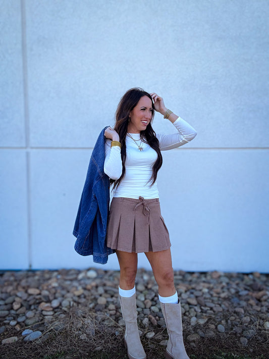 Women wearing a plaid pleated skort with a white long sleeve top and a denim shacket. Paired with tall boots and boot socks 