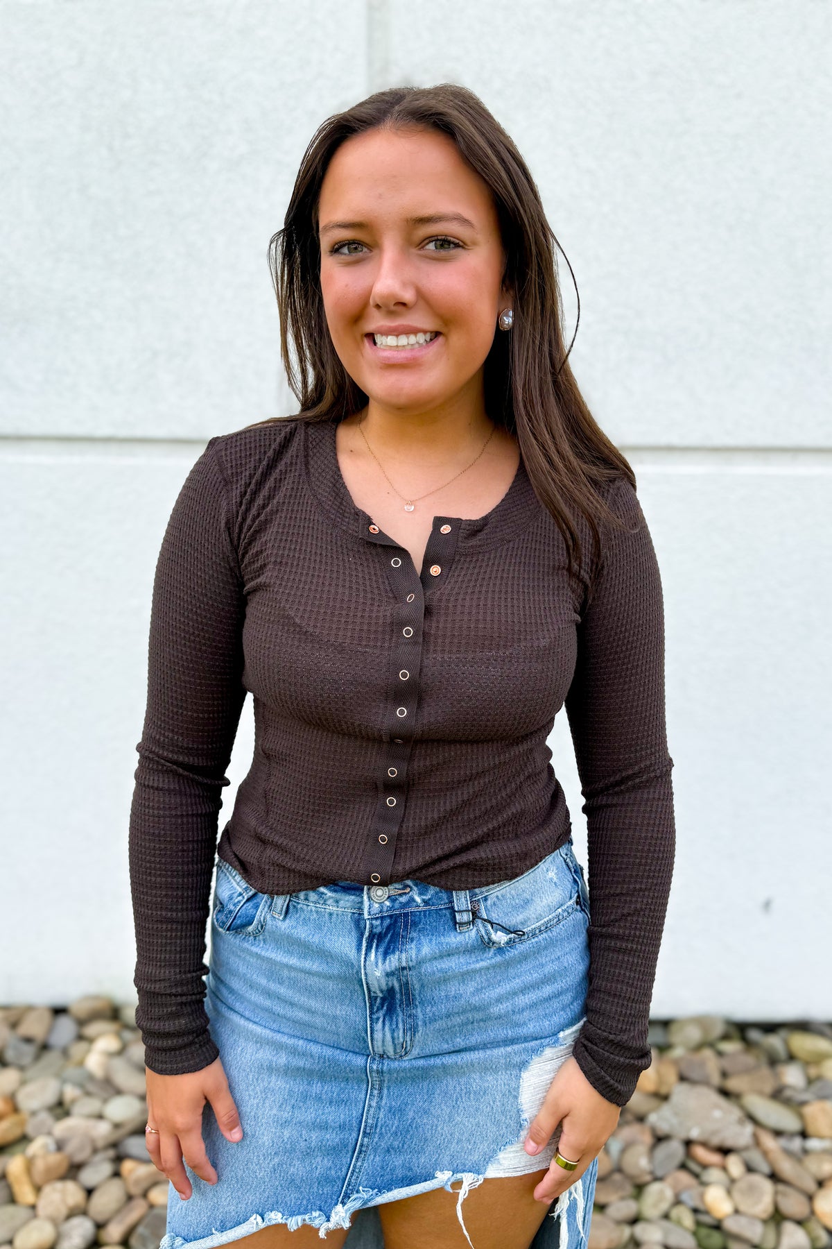 A woman wearing a dark brown long-sleeve waffle-knit henley top and a light blue denim mini skirt, standing outdoors against a light gray wall with pebbles at the bottom.