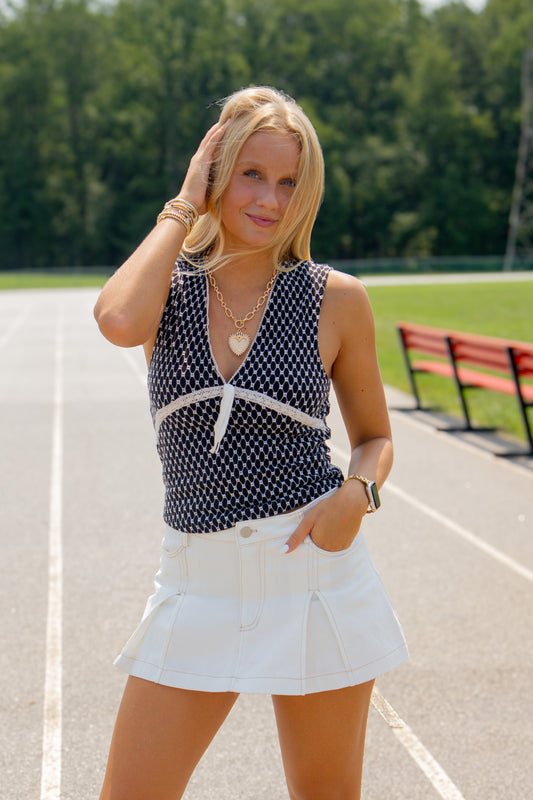 A woman posing on an outdoor athletic track wearing a navy blue and white polka dot sleeveless top with a white tie detail at the neckline, paired with a white pleated mini skirt.