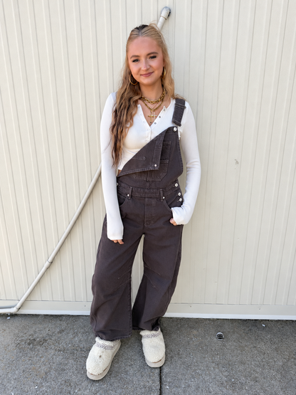 Model wearing the Free People Good Luck Barrel Overalls in Chocolate Mousse, featuring a relaxed wide barrel-leg silhouette, adjustable straps, front bib pocket, and side button detailing, styled with a white ribbed long-sleeve top and cream fuzzy clogs.
