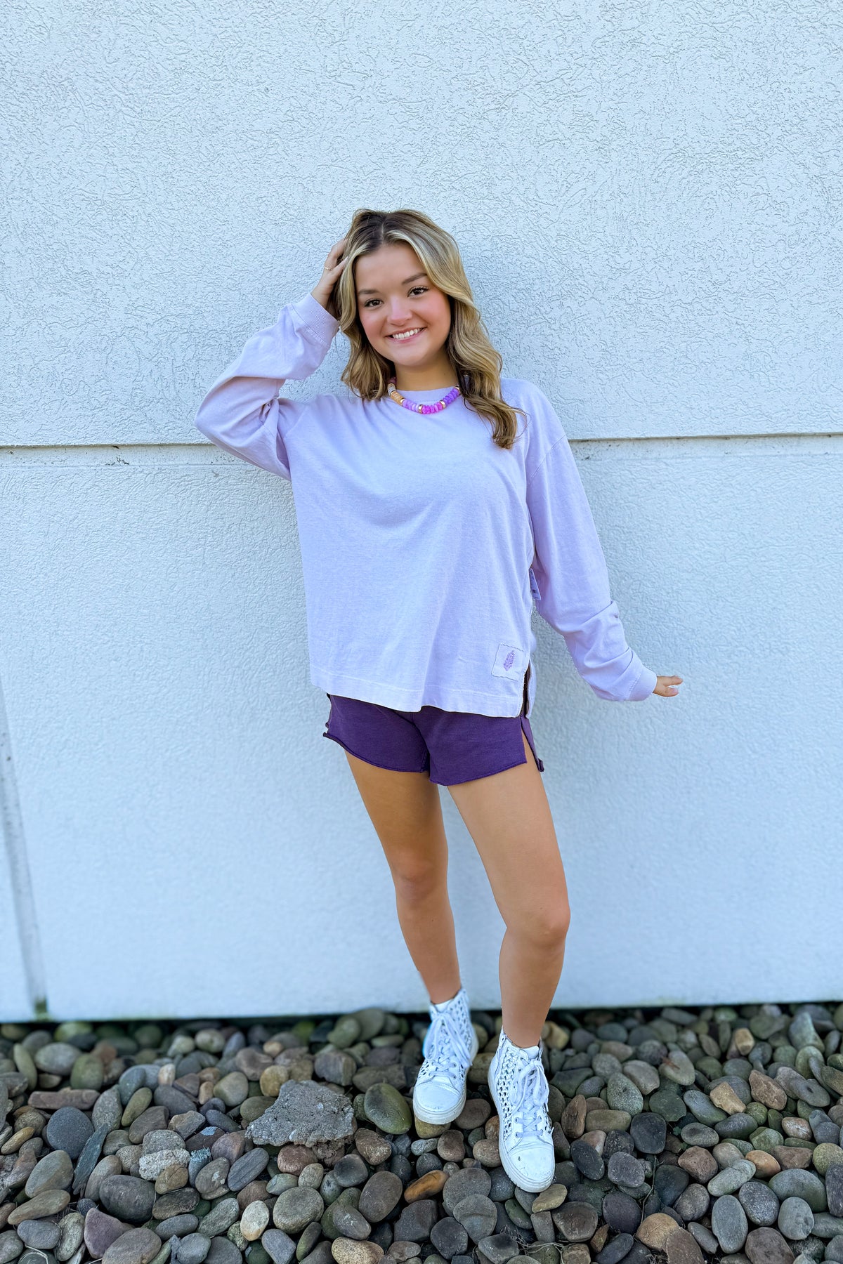 A young woman stands against a white stucco wall on a bed of small gray rocks, wearing a light lavender long-sleeve crewneck shirt, dark purple athletic shorts, white socks, and white sneakers.