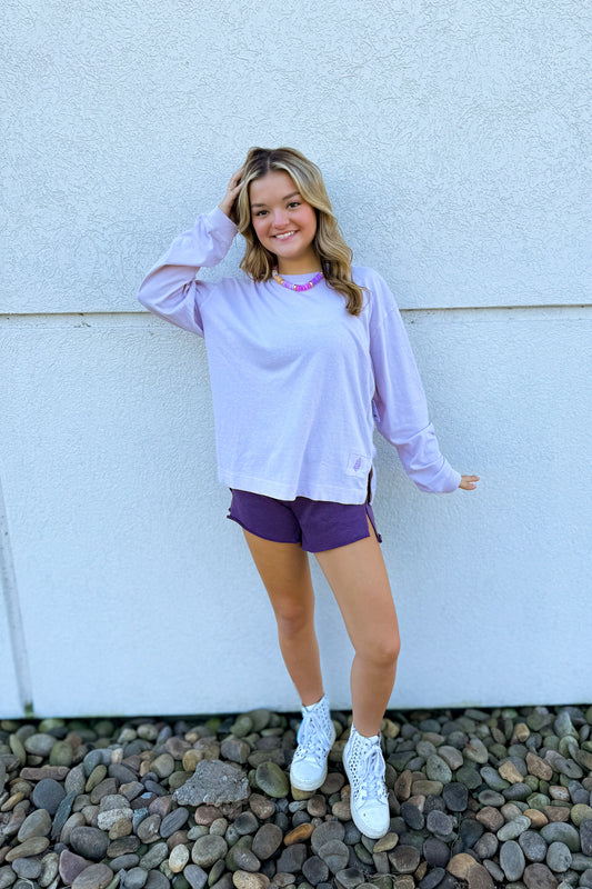 A young woman stands against a white stucco wall on a bed of small gray rocks, wearing a light lavender long-sleeve crewneck shirt, dark purple athletic shorts, white socks, and white sneakers.