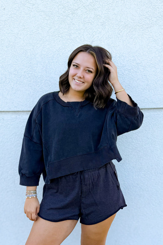 A woman wearing a matching black lounge set consisting of a cropped long-sleeve top and shorts, posing against a light blue textured wall.
