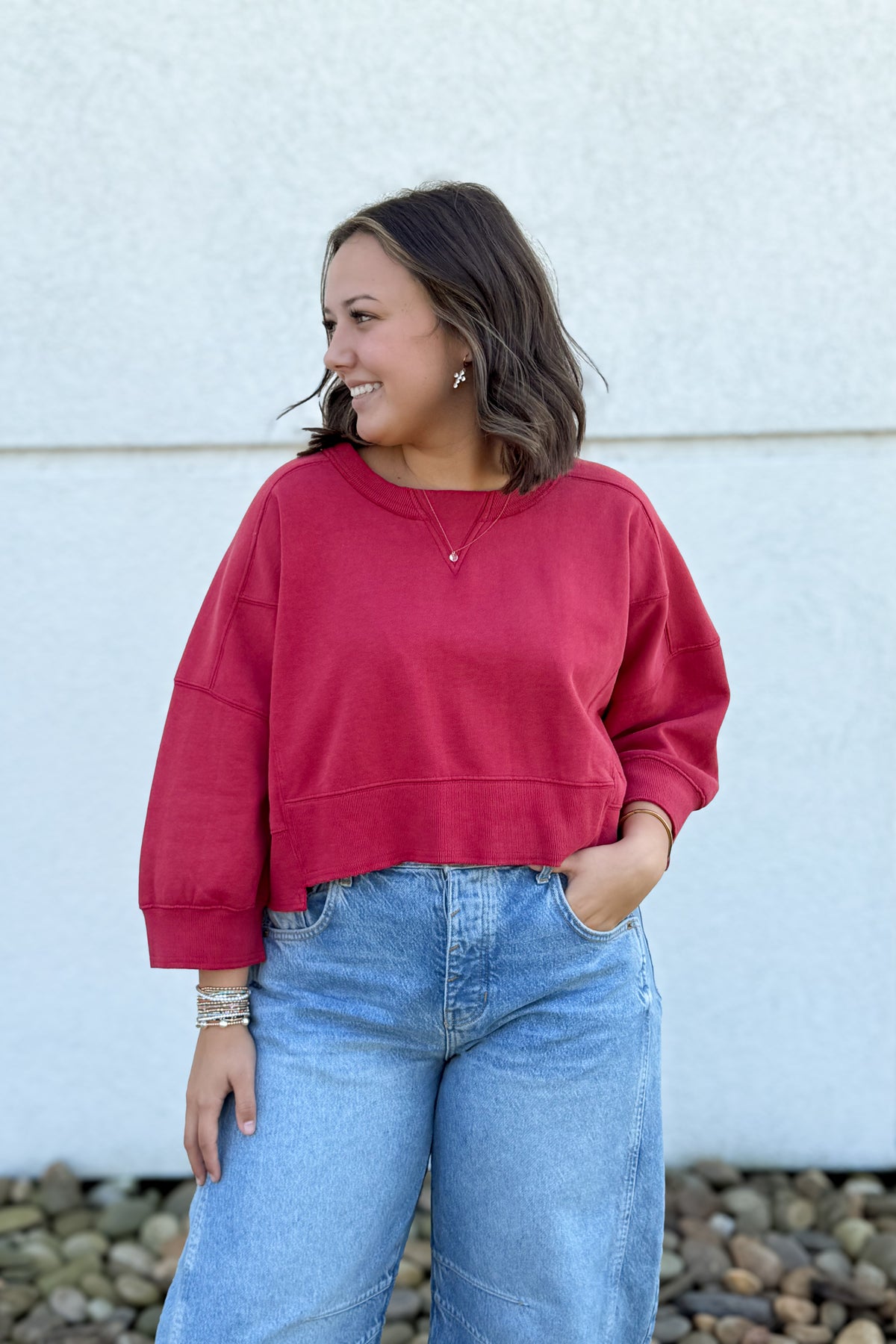 A woman wearing a dark red cropped crewneck sweatshirt and light blue jeans, standing outdoors against a light-colored stucco wall with pebbles at the bottom.