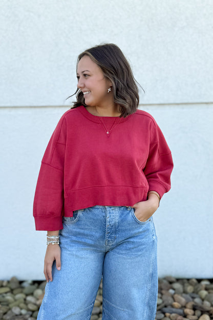 A woman wearing a dark red cropped crewneck sweatshirt and light blue jeans, standing outdoors against a light-colored stucco wall with pebbles at the bottom.