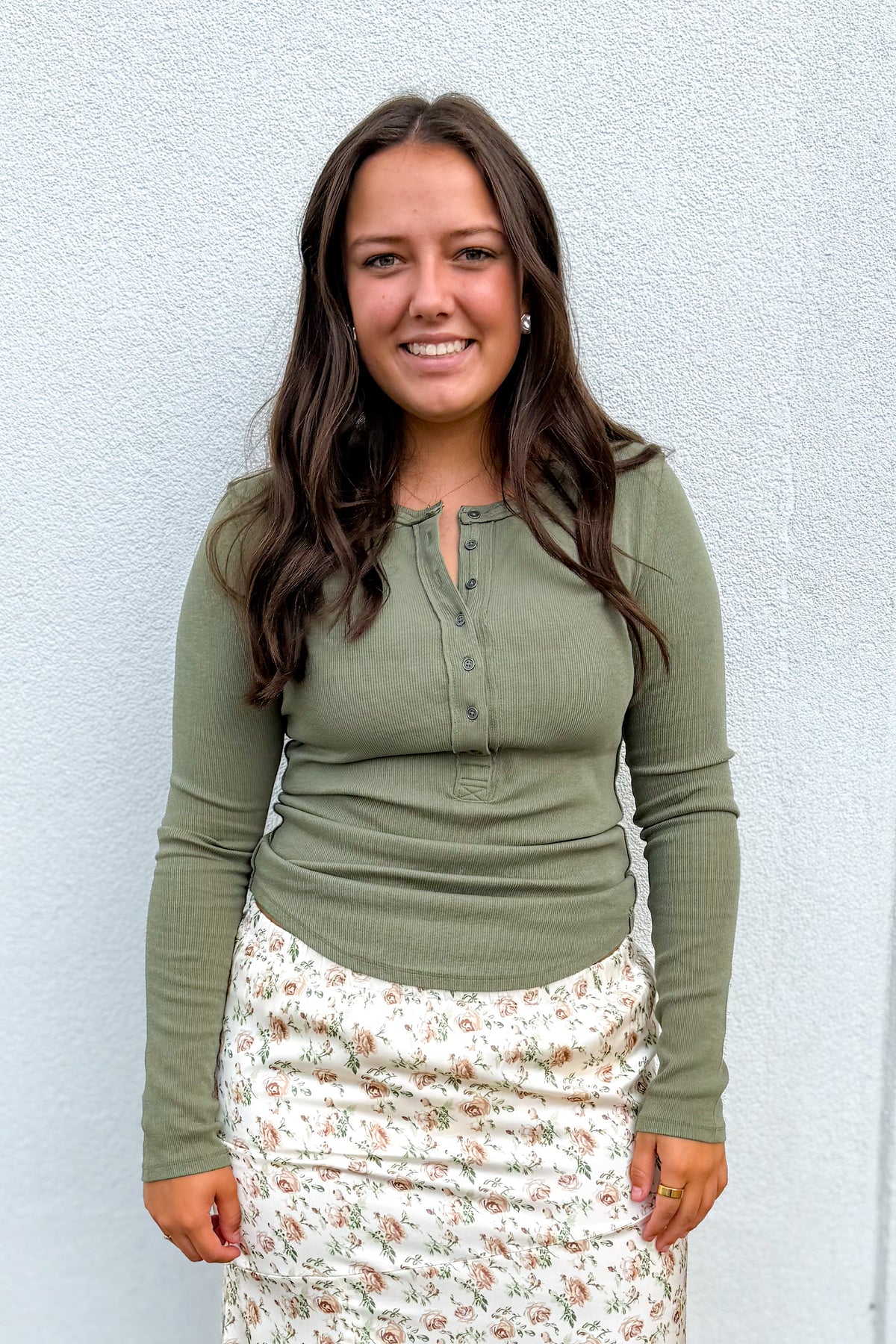 A woman with long brown hair smiling, wearing an olive green long-sleeve henley top and a cream floral skirt, standing against a light gray textured wall.