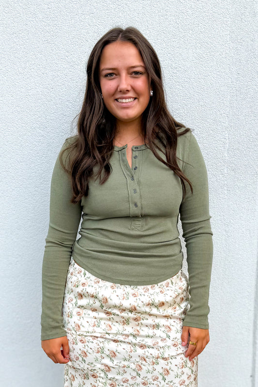 A woman with long brown hair smiling, wearing an olive green long-sleeve henley top and a cream floral skirt, standing against a light gray textured wall.