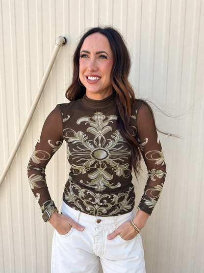Model wearing the Free People Jessa Long Sleeve Top in Olive Combo, featuring a sheer mesh fabric, fitted silhouette, mock neckline, and vintage-inspired olive and cream scroll print, styled with white denim jeans.