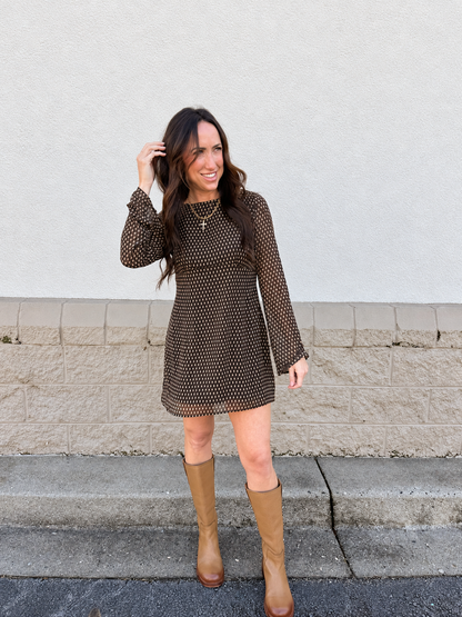 Model wearing the Free People Jolie Printed Mini Dress in Dark Black Brown Combo, featuring a vintage-inspired print, flattering empire waist, sheer long sleeves, and a flared mini silhouette, styled with tan knee-high boots for a chic fall outfit.