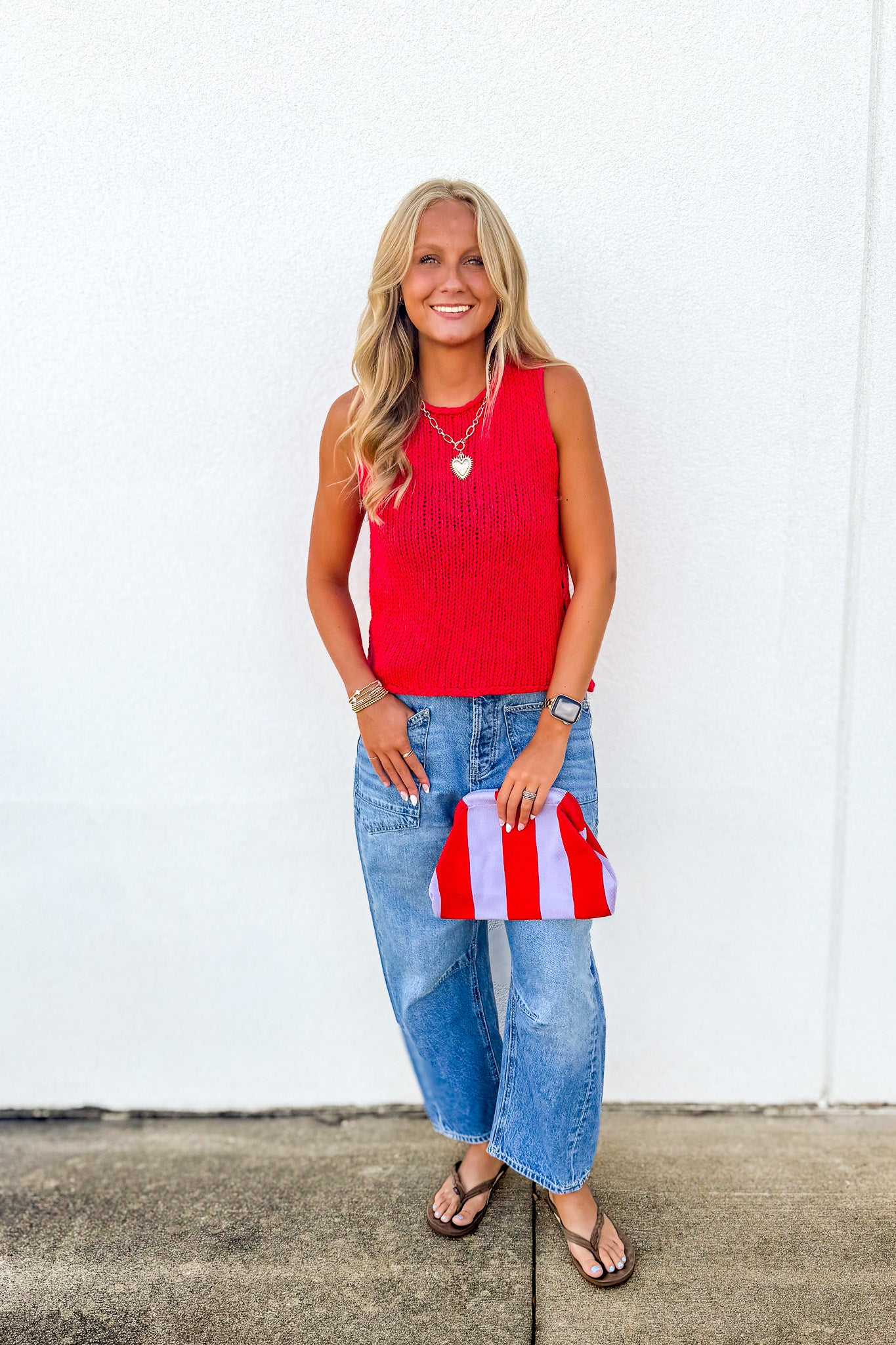 A woman stands against a white wall wearing a bright red sleeveless knit top, loose light-wash blue jeans, and brown sandals, while holding a small red-and-white striped clutch bag.