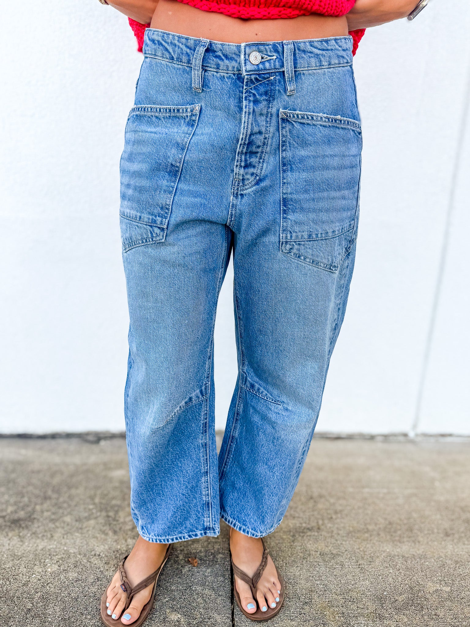 A person wearing light blue high-waisted cropped denim jeans with front patch pockets, paired with brown thong sandals and a red top, standing against a white wall.