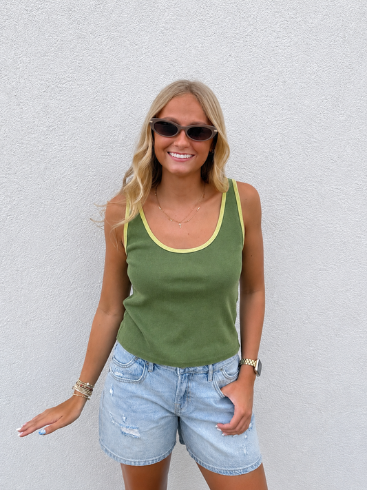 Woman modeling an olive green ribbed contrast trim tank top with light wash distressed denim shorts. Casual summer boutique outfit styled with layered gold necklaces, sunglasses, bracelets, and a gold watch, photographed outdoors against a white textured wall. Perfect everyday warm-weather look.
