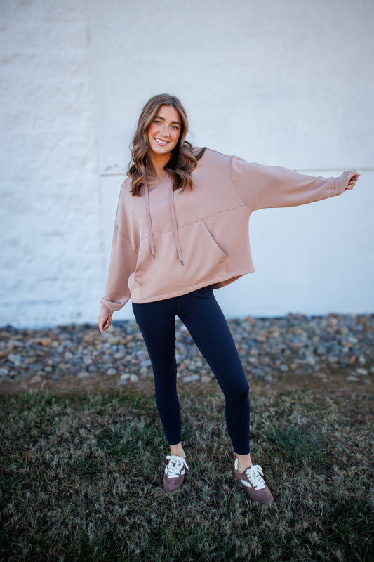Woman wearing a beige hoodie and dark leggings standing against a light-colored wall.