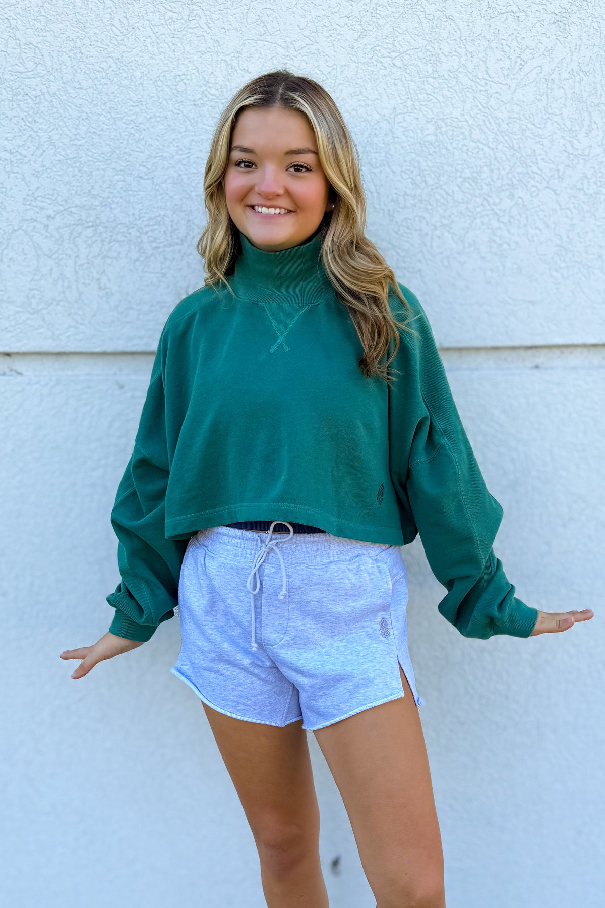 A young woman smiling and posing in front of a textured white wall, wearing a dark green long-sleeve mock neck sweatshirt and light blue heathered athletic shorts.