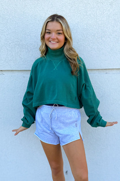 A young woman smiling and posing in front of a textured white wall, wearing a dark green long-sleeve mock neck sweatshirt and light blue heathered athletic shorts.