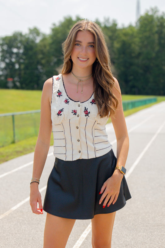 Model wearing a cream knit button-front tank with garnet and black embroidered floral details, paired with a black skort. Styled with layered gold necklaces and a gold watch, this game day outfit is photographed on an outdoor track, making it the perfect look for South Carolina football season.