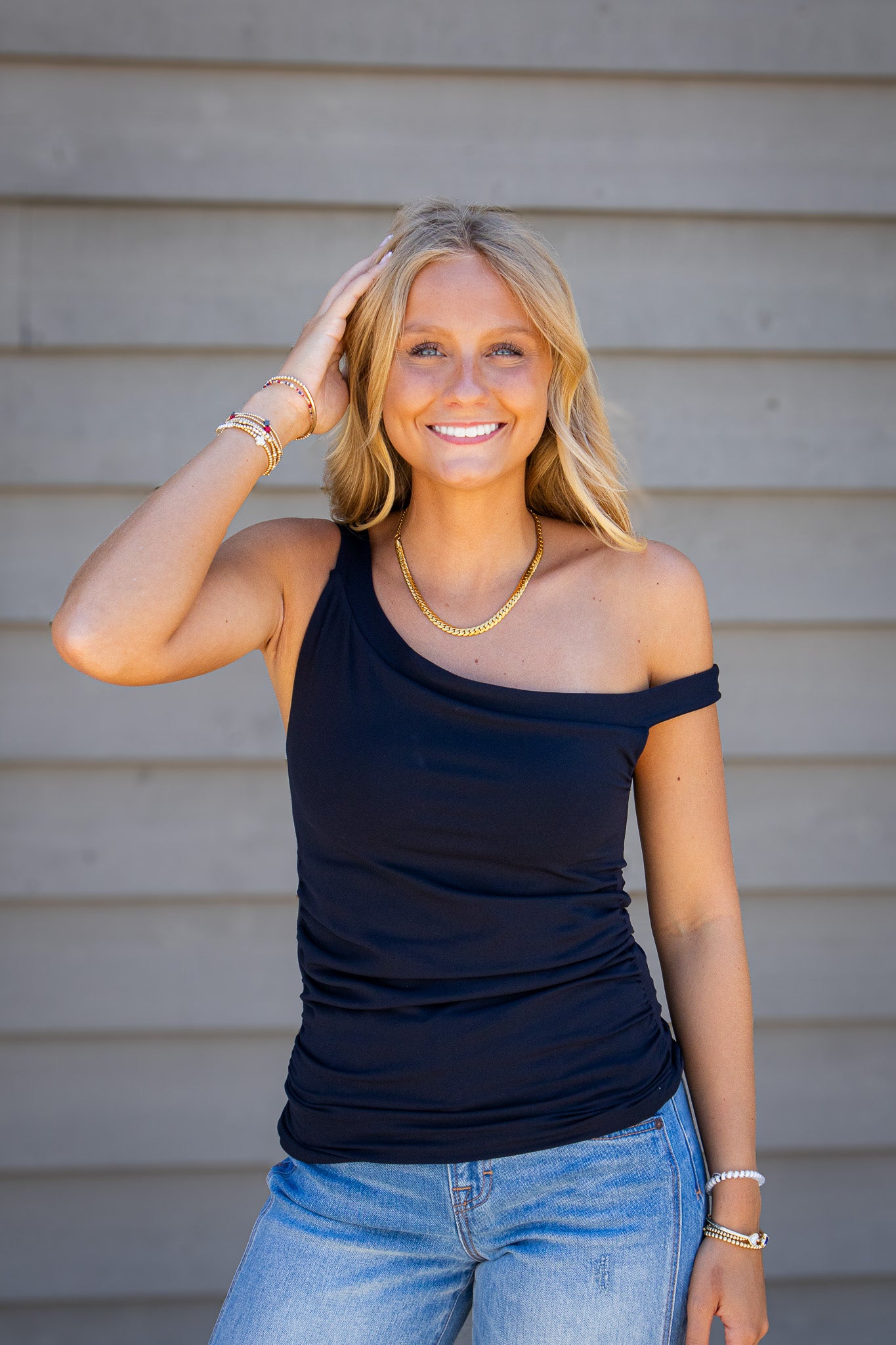 Woman wearing a fitted black one-shoulder top with ruched side detailing and light wash jeans, styled with layered gold jewelry and bracelets, standing outdoors against a neutral background.