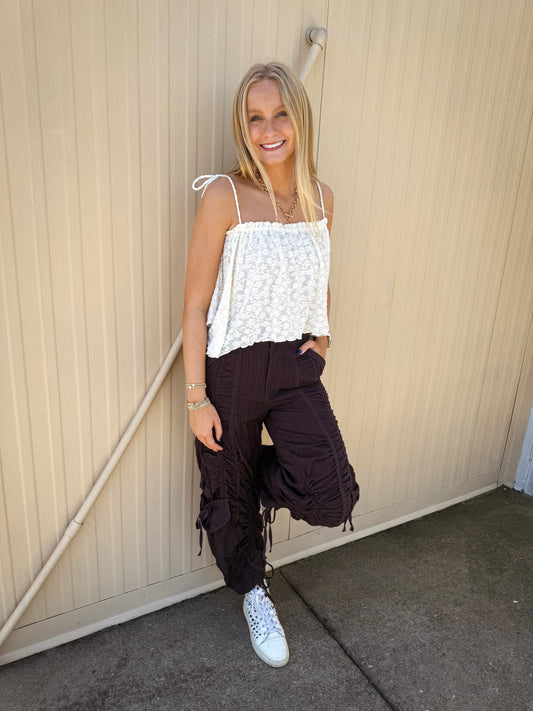Model wearing Free People Scorpio Shirred Mid-Rise Cargo Pants in Black Coffee, featuring adjustable shirred drawstring detailing, cargo pockets, a relaxed mid-rise fit, and utility-inspired styling paired with a white textured cami and white sneakers.