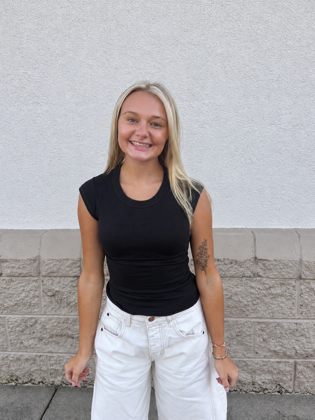 Model wearing the Free People So Soft Seamless Modal Baby Tee in Black, featuring a fitted ribbed design, crew neckline, cap sleeves, and waist-length silhouette, styled with white denim jeans for an effortless everyday look.