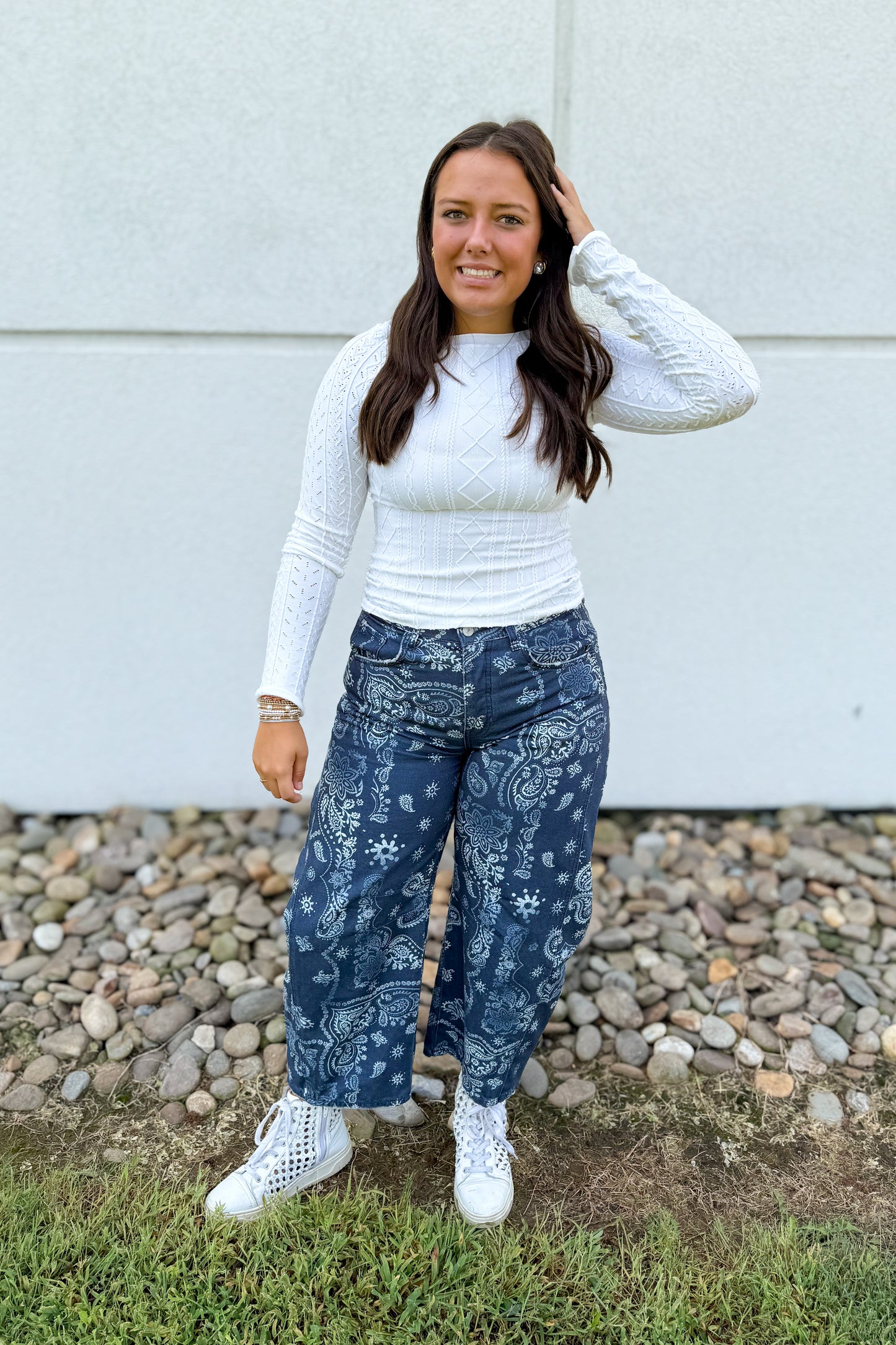 A woman stands outdoors in front of a light gray wall and gravel, wearing a white textured long-sleeve top, dark blue wide-leg jeans with a white paisley and floral pattern, and white sneakers.