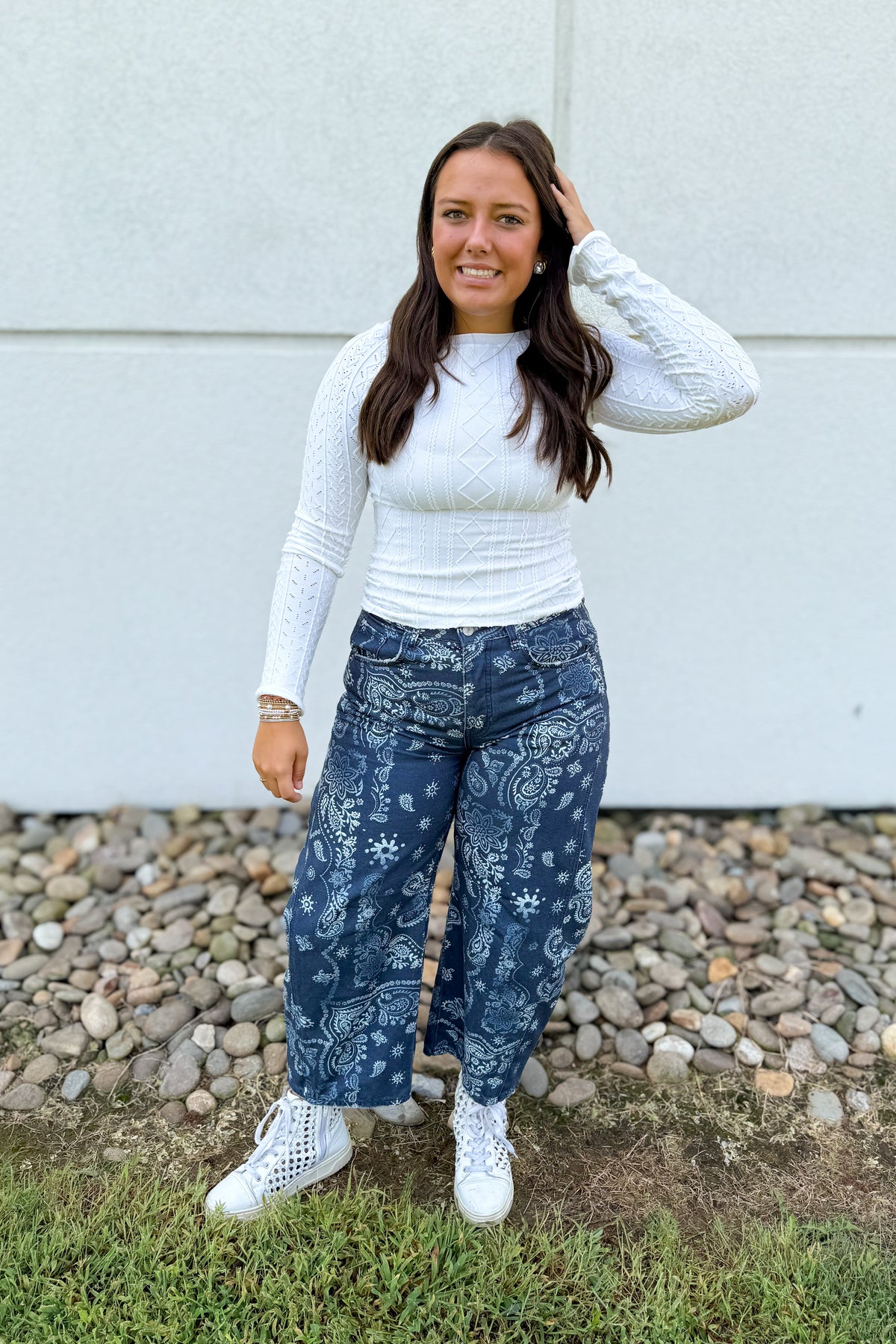 A woman stands outdoors in front of a light gray wall and gravel, wearing a white textured long-sleeve top, dark blue wide-leg jeans with a white paisley and floral pattern, and white sneakers.