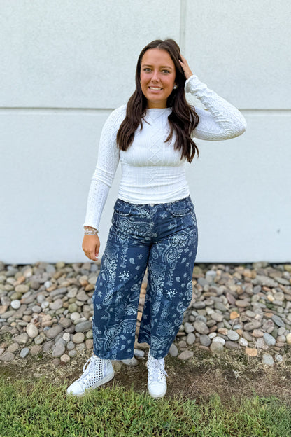 A woman stands outdoors in front of a light gray wall and gravel, wearing a white textured long-sleeve top, dark blue wide-leg jeans with a white paisley and floral pattern, and white sneakers.