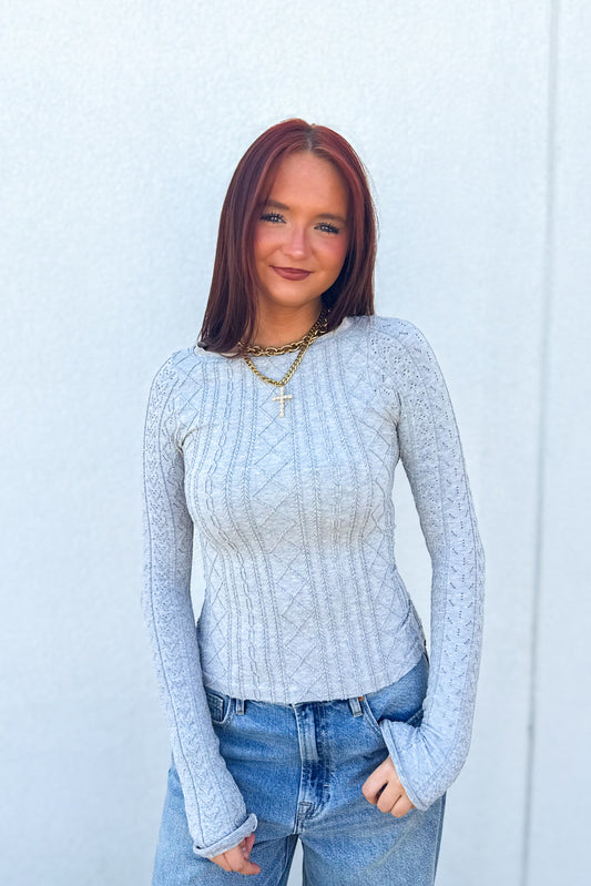A woman with reddish-brown hair wearing a light-gray cable-knit long-sleeve top and blue jeans, standing against a light gray textured wall.