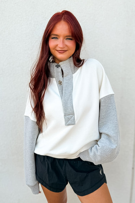 Model wearing an ivory and heather gray colorblock pullover with a high collar, oversized snap-button placket, long sleeves, and a relaxed fit, styled with black athletic shorts.