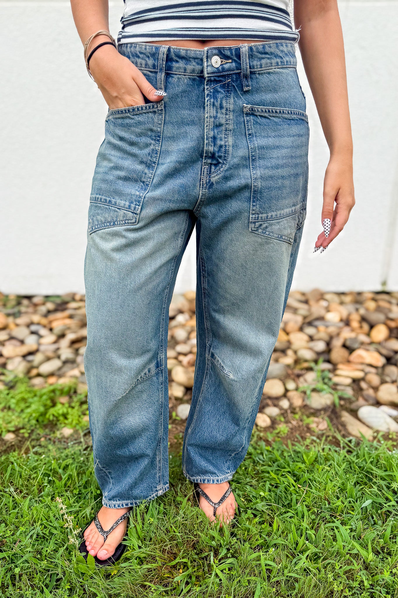 A person wearing light blue high-waisted denim jeans with patch pockets, standing outdoors on grass with a pebble bed and white wall in the background.