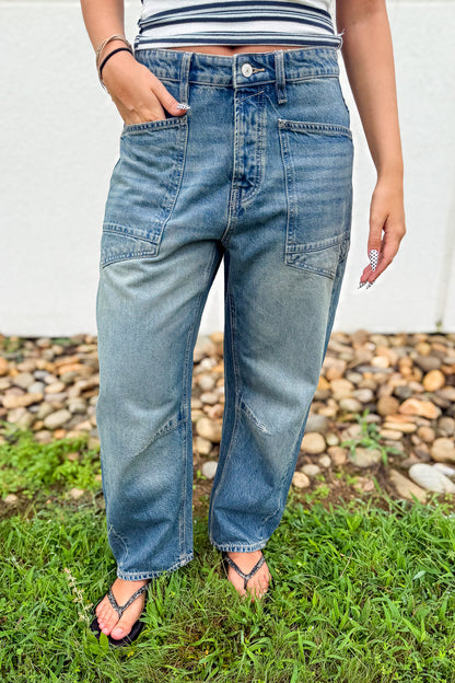 A person wearing light blue high-waisted denim jeans with patch pockets, standing outdoors on grass with a pebble bed and white wall in the background.