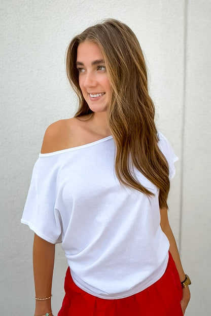 Model wearing an white Free People We The Free oversized off-the-shoulder tee with a relaxed draped fit and short sleeves, styled with red shorts.