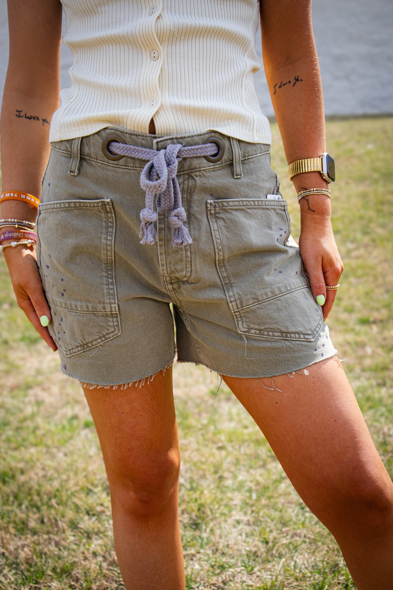 Olive green denim shorts with oversized front pockets, raw hem detail, and rope drawstring waist, styled with a cream ribbed tank top for a casual summer outfit outdoors
